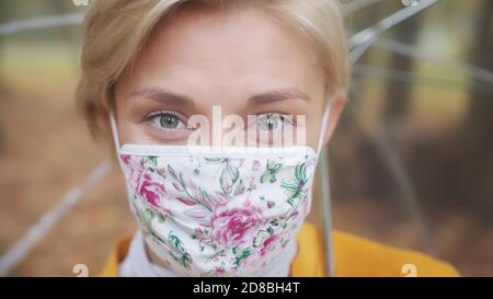 Giovane donna bionda con maschera facciale alla moda e cappotto giallo che tiene ombrello tranparetn nel parco. Foto di alta qualità Foto Stock
