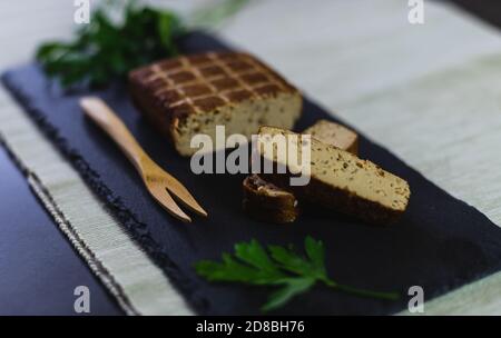Un pezzo di nutriente tempeh di soia tagliato a fette su un piatto di ardesia con prezzemolo fresco e una forchetta di legno. Foto Stock