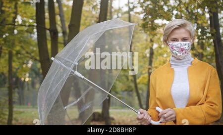 Giovane caucasica donna alla moda con maschera facciale che gira umbrela trasparente nel parco in autunno. . Foto di alta qualità Foto Stock