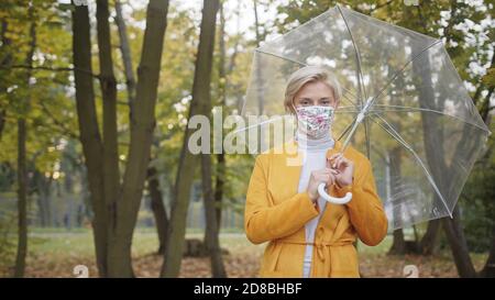 Bella donna caucasica con maschera facciale tenuta con umbrela trasparente nel parco in autunno. Foto di alta qualità Foto Stock