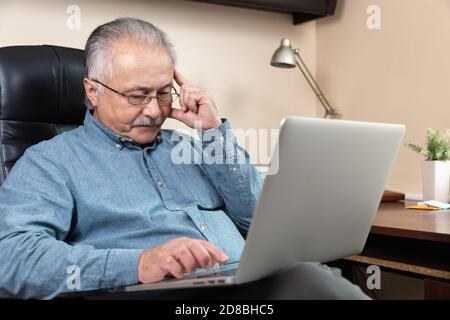 Pensiva uomo d'affari senior lavoro a casa. Un uomo anziano in occhiali sta lavorando a distanza utilizzando un computer portatile. Lavoro remoto durante il concetto di coronovirus Foto Stock