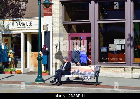 Traverse City, Michigan, Stati Uniti. Una coppia che indossa le maschere del viso durante la pandemia del coronavirus passa una donna seduta su una panchina lungo Front Street. Foto Stock