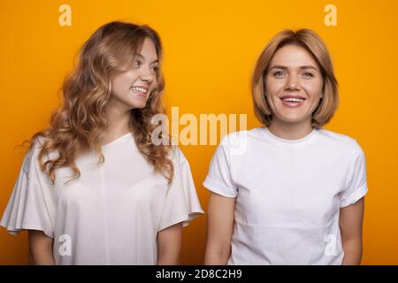 Donna caucasica con capelli biondi che la guarda sorridere sorella in t-shirt bianca su una parete gialla dello studio Foto Stock