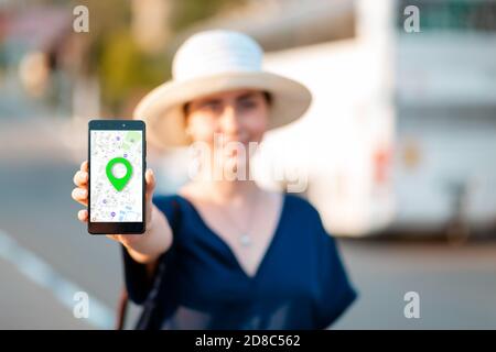 Una donna in cappello di paglia tiene un cellulare con un'app mappa online con sfondo mark, sfocato. Sullo sfondo c'è una strada e un autobus. Il concetto di n on-line Foto Stock