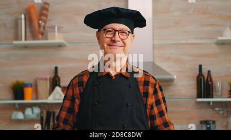 Vecchio uomo caucasico che indossa grembiule in cucina casalinga sorridendo alla macchina fotografica. Panettiere anziano in pensione con bonete preparazione di ingredienti di pasticceria su tavola di legno pronto a cucinare pane, torte e pasta gustosi fatti in casa. Foto Stock