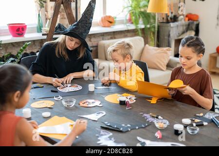 Concentrato giovane insegnante d'arte in streghe cappello seduto a tavola E disegno Halloween immagini con i capretti mentre preparando le decorazioni di carta