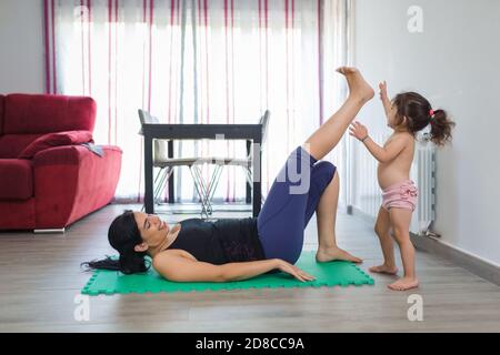 La madre latino pratica le posizioni di yoga e di idoneità a casa su un tappetino con la sua bambina di due anni sul pavimento della sala da pranzo. Foto Stock