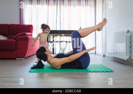 La madre latino pratica le posizioni di yoga e di idoneità a casa su un tappetino con la sua bambina di due anni sul pavimento della sala da pranzo. Foto Stock