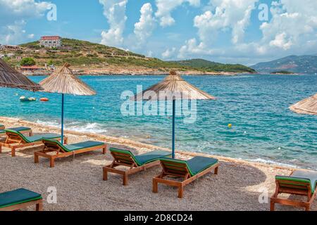 Vista della spiaggia vuota - sdraio e ombrelloni vicino all'acqua di mare, ciottoli e sabbia dorata, montagne all'orizzonte e nuvole sul cielo. Paesaggio di aba Foto Stock