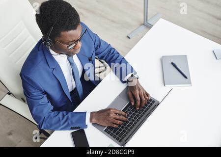 Vista ad alto angolo di un uomo d'affari africano in cuffia che digita laptop e comunicazione online sul suo posto di lavoro in ufficio Foto Stock