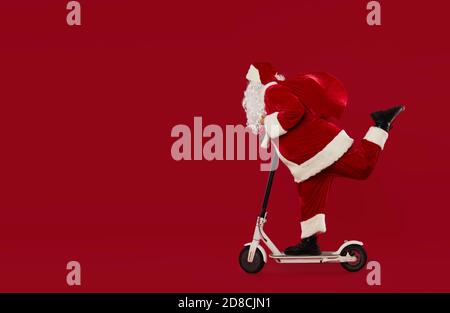 Ritratto a lunghezza intera di babbo natale in sella a uno scooter elettrico isolato su sfondo rosso. Concetto di consegna rapida per le festività Foto Stock