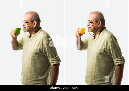Indiano asiatico anziano o anziano che beve succo d'arancia fresco in un bicchiere mentre si è in piedi su sfondo bianco Foto Stock