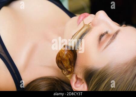 Una grande lumaca Akhatina striscia sul volto di una giovane donna e ringiovanisce la sua pelle. Il concetto di cura insolita e ringiovanimento della pelle del viso. Foto Stock