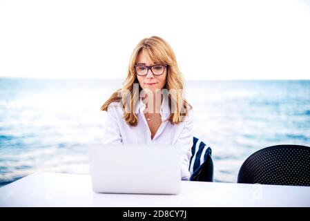Un'immagine di donna matura sorridente seduta al tavolo con il computer portatile sullo sfondo del mare. Foto Stock
