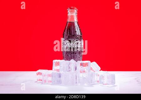 Londra, Regno Unito - Ottobre 29 2020: Una bottiglia di Coca Cola di vetro ghiacciato si trova in un mucchio di cubetti di ghiaccio con condensa su sfondo rosso. Foto Stock