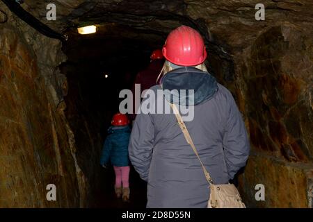 primo piano di donna che indossa un casco rosso protettivo e una giacca grigia e un bambino piccolo con casco in lontananza, all'interno di una grotta scura Foto Stock