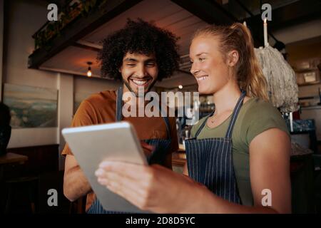 Ritratto di colleghi di sesso maschile e femminile che indossano grembiule e ridono durante la visione di video in un tablet digitale al bar Foto Stock