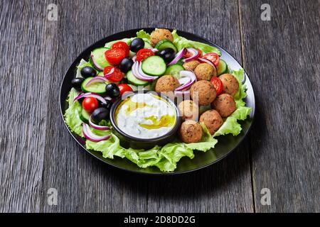 Piatto mediterraneo di falafel, pomodori, cetrioli, cipolla rossa, olive nere, sulle foglie fresche di insalata servite con salsa tzatziki servita su un nero Foto Stock