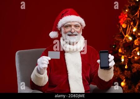 Ritratto dell'uomo in costume di Babbo Natale con biglietto da visita e il telefono cellulare in mano per l'annuncio per le vacanze Foto Stock
