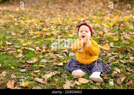 Baby girl in giacca gialla e archetto rosso seduta sull'erba, giocando nelle foglie autunnali. Foto Stock