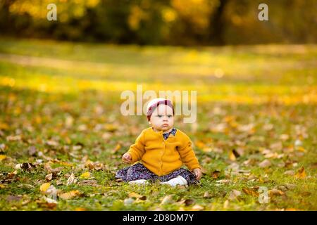 Baby girl in giacca gialla e archetto rosso seduta sull'erba, giocando nelle foglie autunnali. Foto Stock