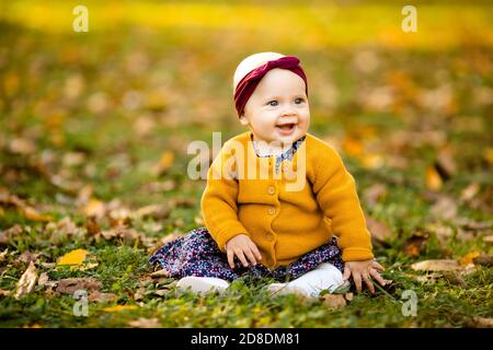 Baby girl in giacca gialla e archetto rosso seduta sull'erba, giocando nelle foglie autunnali. Foto Stock