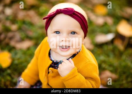 Baby girl in giacca gialla e archetto rosso seduta sull'erba, giocando nelle foglie autunnali. Foto Stock