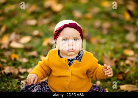 Baby girl in giacca gialla e archetto rosso seduta sull'erba, giocando nelle foglie autunnali. Foto Stock