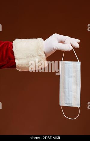 Primo piano di uomo in costume di Santa che tiene maschera protettiva durante pandemia su sfondo marrone Foto Stock