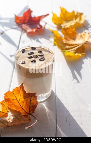 Bevanda tradizionale coreana al latte caffeinato, caffè Dalgona in un bicchiere trasparente con chicchi di caffè su sfondo bianco. Foglie di acero secche. Autu Foto Stock