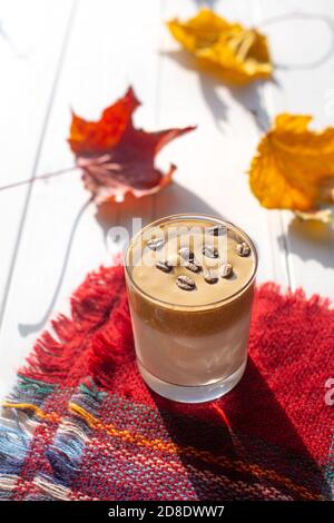 Bevanda tradizionale coreana al latte caffeinato, caffè Dalgona in un bicchiere con chicchi di caffè su sfondo bianco. Foglie di acero secche. A scacchi caldi Foto Stock