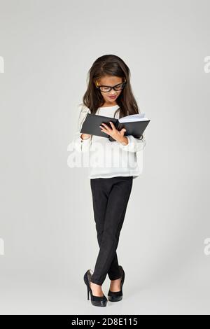 carino bambina ragazza in oversize le scarpe e gli occhiali da mamma leggere notebook su sfondo bianco. bambino che gioca donna d'affari Foto Stock