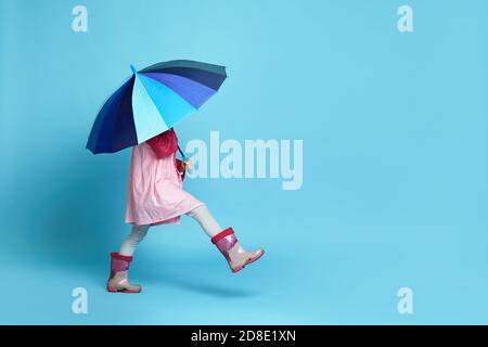 felice bambina con ombrello blu in giacca rosa e stivali in gomma su sfondo blu. copia spazio per il testo Foto Stock