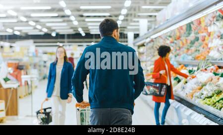 Al supermercato: Uomo che spinge il Carrello della spesa attraverso la sezione prodotti freschi del negozio. Negozio con molti clienti Shopping. Seguito dello scatto della vista posteriore Foto Stock