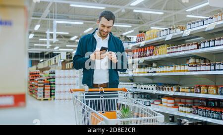 Al supermercato: L'uomo bello usa smartphone, sorrisi e si leva con il carrello della spesa nella sezione delle merci in scatola. Foto Stock