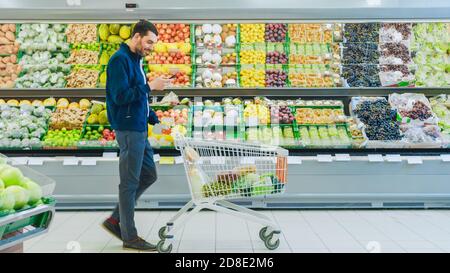 Al supermercato: Bell'uomo con smartphone, spinge il carrello della spesa, cammina oltre la sezione prodotti freschi del negozio. Uomo immerso in Internet Foto Stock