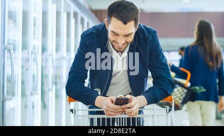 Al supermercato: Il bell'uomo usa smartphone e sorrisi, mentre si appoggia sulla Shopping Card. Buon cliente Shopping nella sezione Frozen del Foto Stock