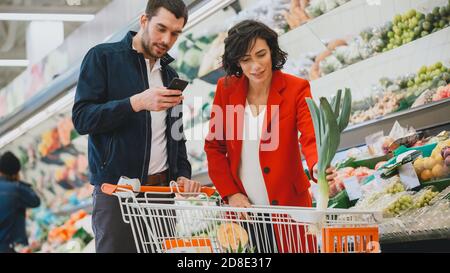 Al supermercato: Happy Couple fa Shopping, scegliendo frutta e verdura nella sezione prodotti freschi. L'uomo usa lo smartphone e spinge lo shopping Foto Stock