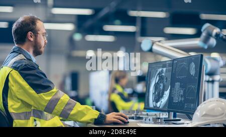 In fabbrica: Ingegnere meccanico lavora su computer, progetta in CAD modello 3D del motore. In background operai e funzionamento della fabbrica Foto Stock