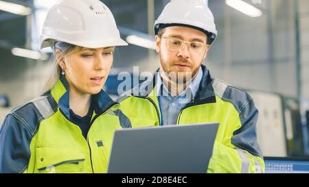 Gli ingegneri industriali maschili e femminili lavorano in uno stabilimento di produzione, discutono progetto, punto nella direzione del macchinario mentre usano i laptop. Foto Stock