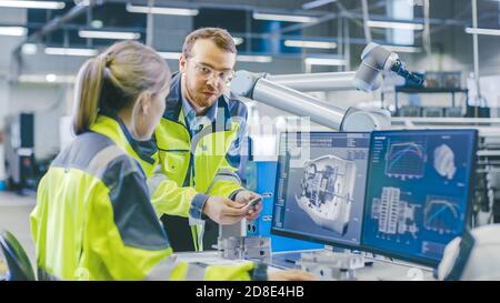 In fabbrica: Il meccanico maschile tiene componenti e il capo ingegnere femminile lavoro su Personal computer, si discute dei dettagli del motore 3D Foto Stock