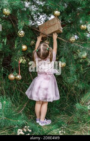 Carino bambina in un vestito rosa sta cercando di ottenere una bella confezione regalo dall'albero di Natale. Nuovo anno e concetto di natale Foto Stock