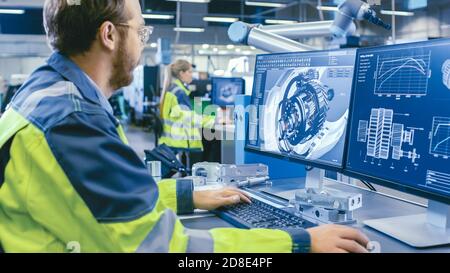 In fabbrica: Ingegnere meccanico lavora su computer, progetta in CAD modello 3D del motore. In background operai e produzione di fabbrica Foto Stock