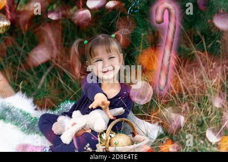 Una bambina carina si siede vicino all'albero di Natale e raggiunge per una bacchetta magica incandescente che si affaccia davanti a lei. Miracolo di Natale. Adempimento dei desideri Foto Stock