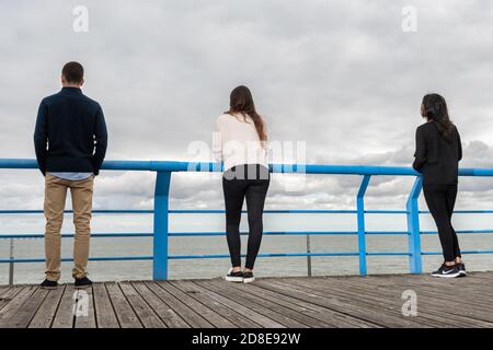 Tre persone si distinguono su una passerella, di fronte all'oceano sotto un cielo nuvoloso, trasmettendo solitudine e contemplazione. Foto Stock