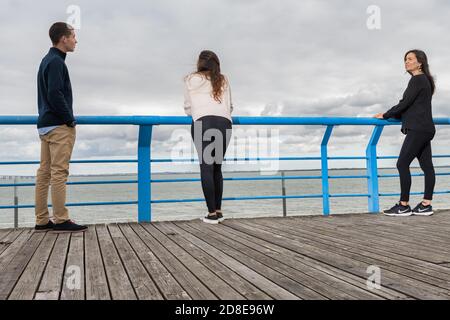 Tre persone si distinguono su una passerella, di fronte all'oceano sotto un cielo nuvoloso, trasmettendo solitudine e contemplazione. Foto Stock