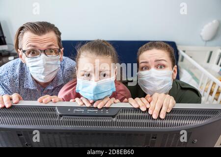Stanco famiglia caucasica indossare maschere mediche a casa, in piedi vicino alla televisione e guardando la macchina fotografica con aspetto incatenato, auto isolamento Foto Stock