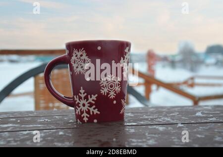 Coppa rossa o borgogna con un modello di fiocchi di neve sullo sfondo di una foresta invernale. Nevicare. Capodanno. Natale. Tazza con una bevanda calda sul Foto Stock