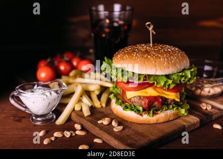 Hamburger fresco e gustoso fatto in casa con lattuga e formaggio su tavola rustica di legno. Patatine fritte, pomodori e salsa. Sfondo di cibo scuro. Foto Stock