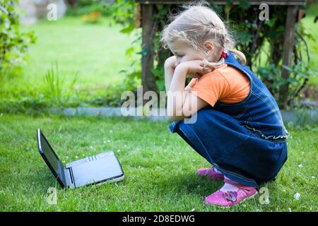 Concentrato bambino guardando cartoni animati con il suo piccolo computer portatile mentre si siede su erba verde in giardino, caucasica ragazza che indossa jeans vestito Foto Stock